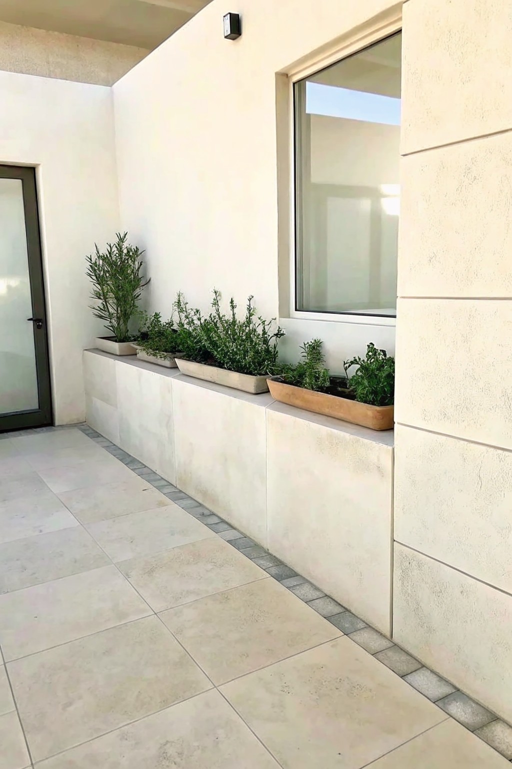 A patio with a long low light-colored ledge holding several planters with green plants beside a large window on a stucco wall.