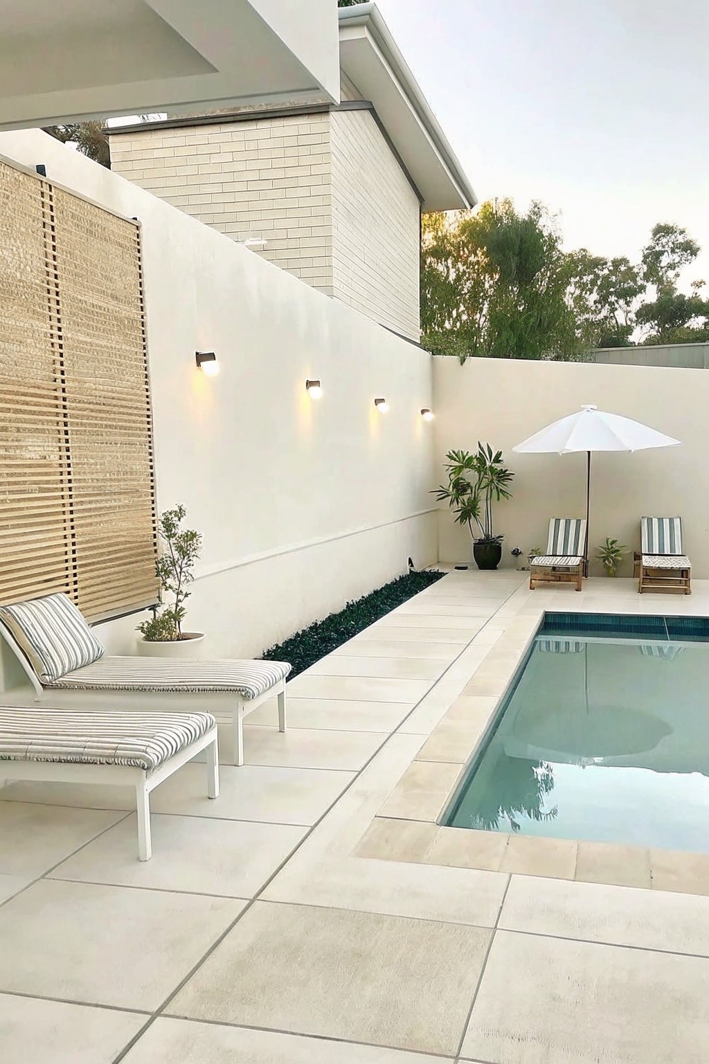 A modern outdoor pool area with light stone paving, a narrow strip of dark green groundcover along a white wall, two striped lounge chairs, a white umbrella, and a rectangular pool.