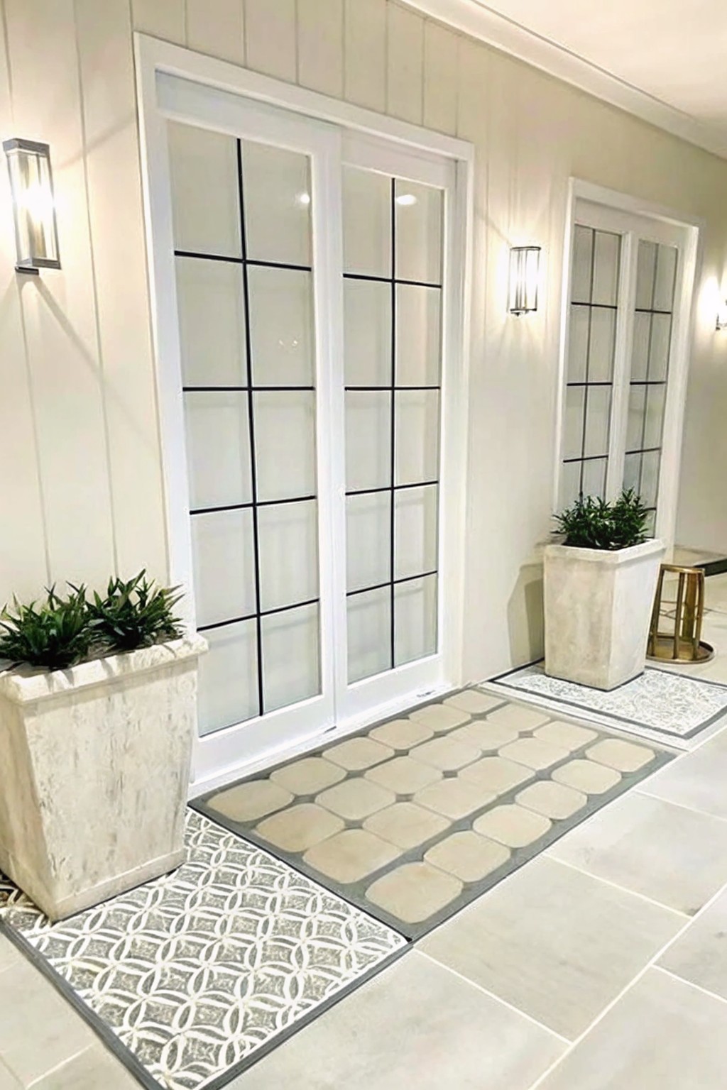 White double doors with black grids, flanked by stone planters and patterned rugs.