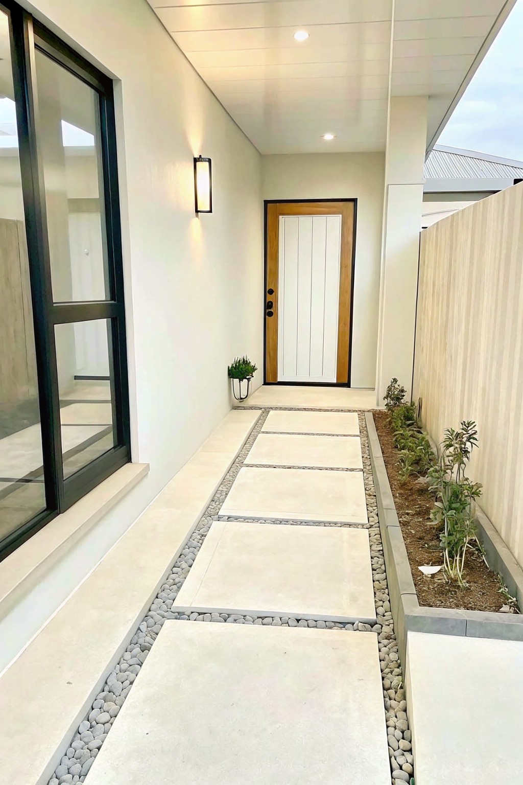 A narrow concrete walkway lined with large square pavers set in gray pebbles, running beside a raised garden bed with young plants and leading to a white door.