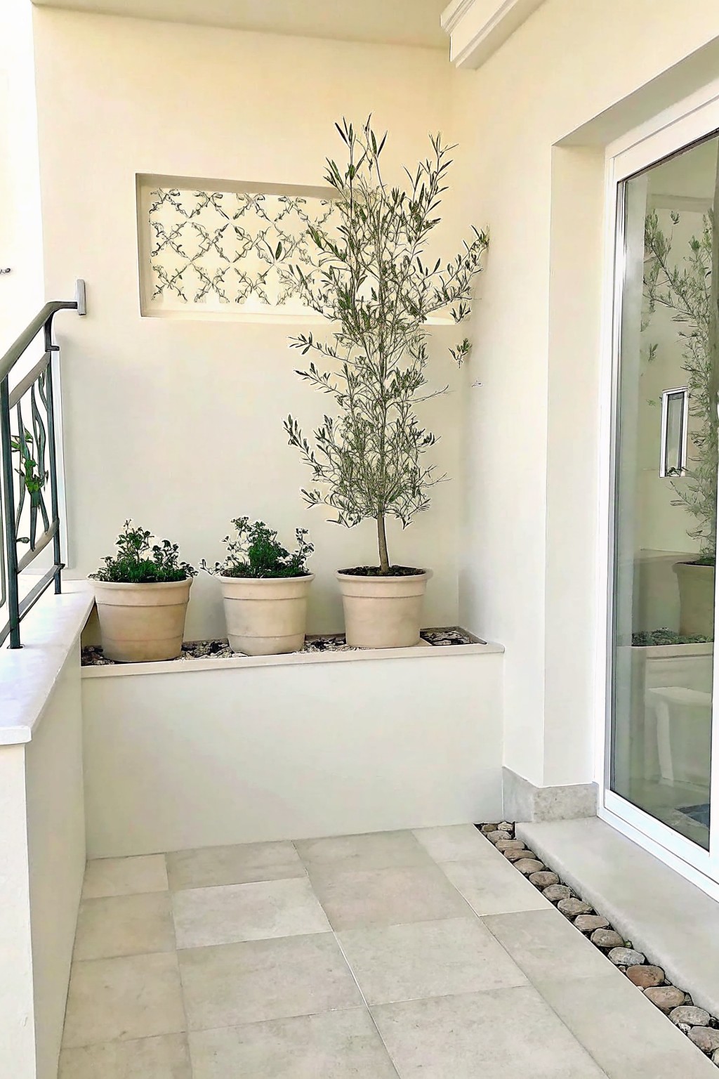 A light-tiled patio with a raised planter holding three pots and a border of small pebbles running along the base of the wall and planter edge.