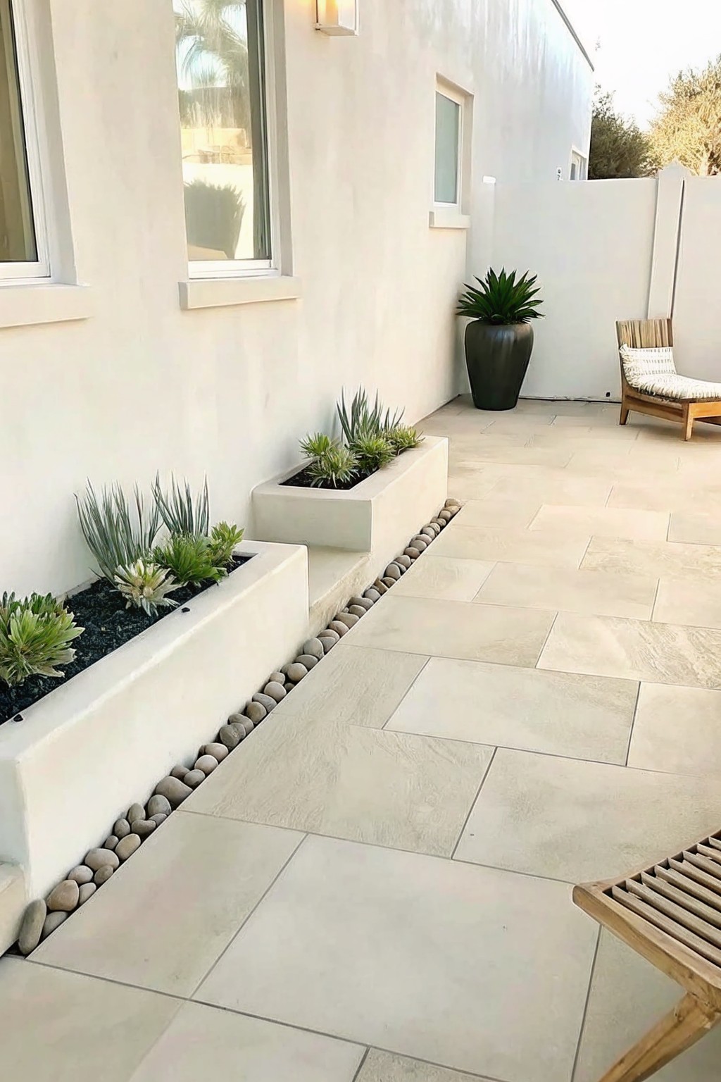A light stone patio with raised rectangular concrete planters filled with succulents, bordered by a line of small gray pebbles next to the paving.