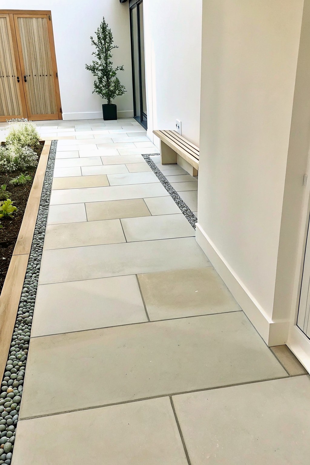 Modern stone walkway with pebble border, wooden bench, potted plant, and garden bed