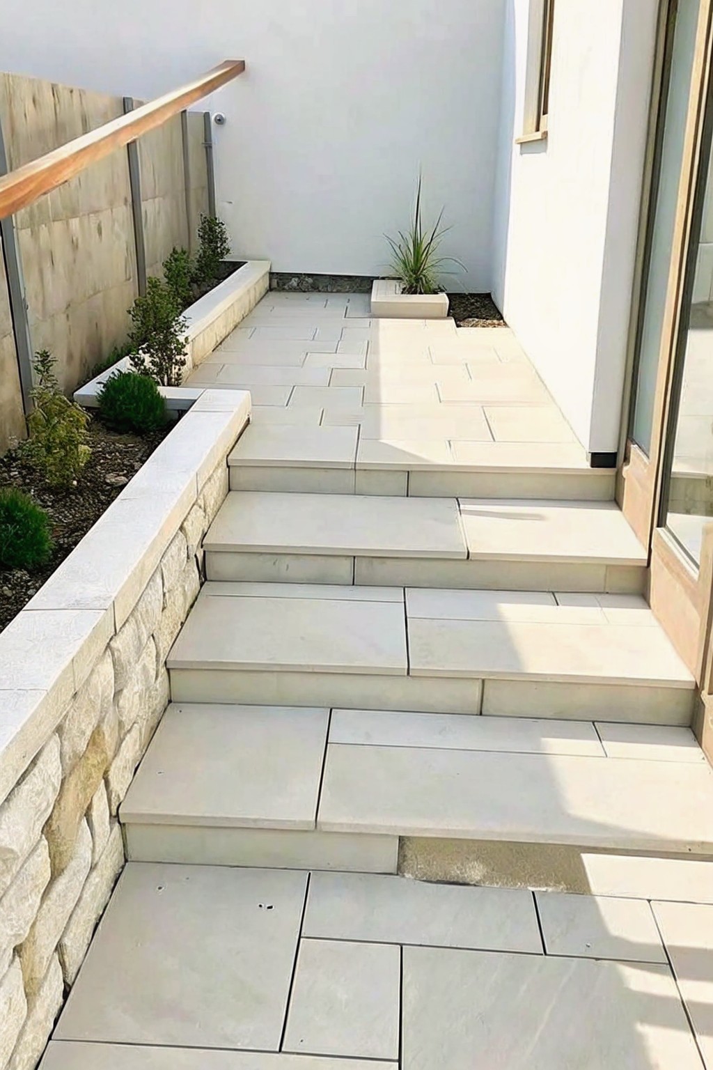 A descending set of light stone paver steps runs beside a low stacked stone wall with narrow planted beds on the left.