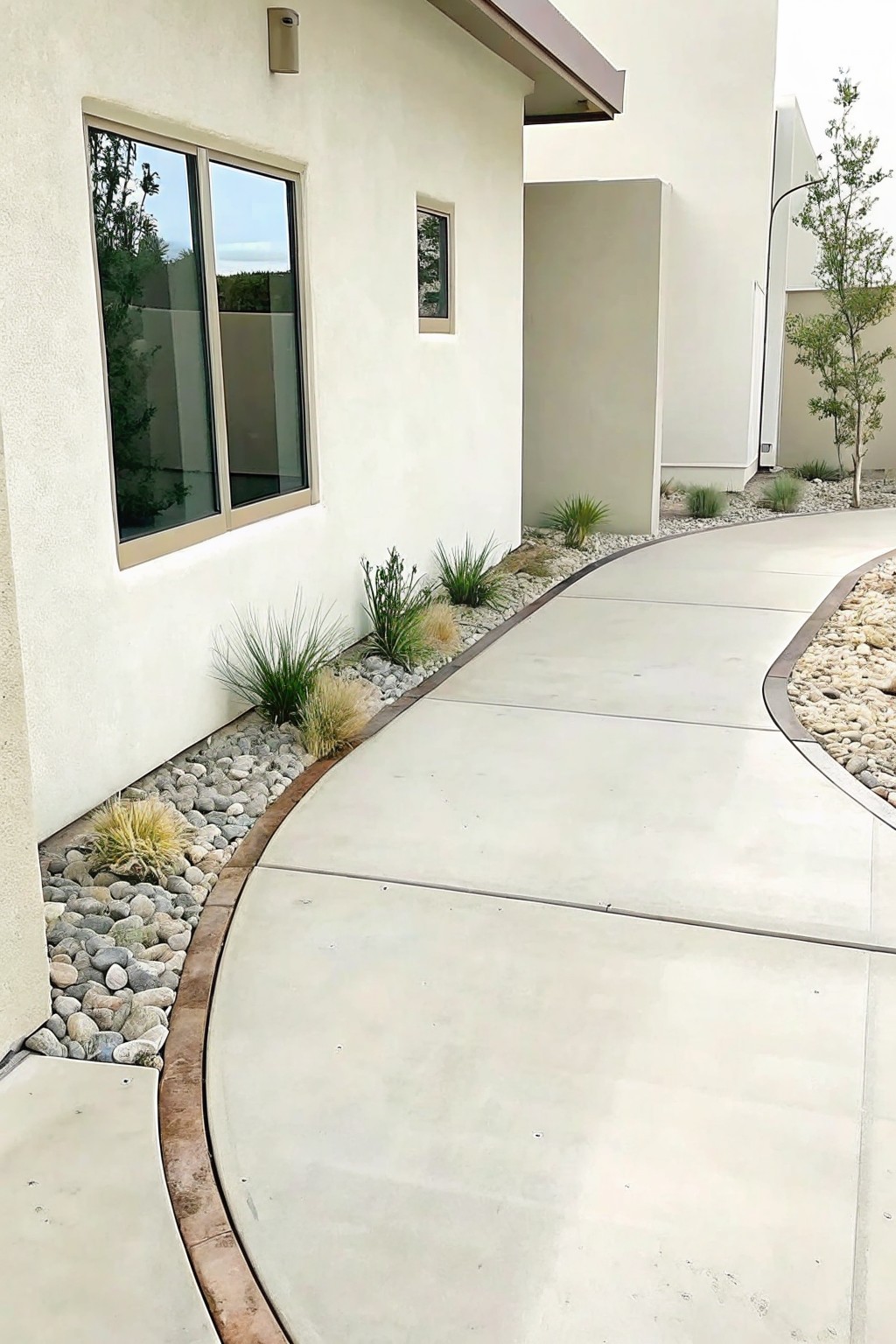 Modern beige house with large window beside curved concrete path and rock landscaping