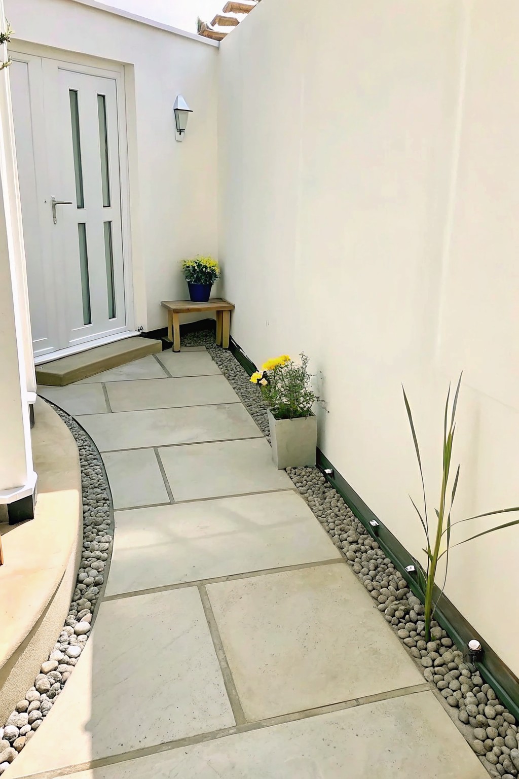 A narrow paved walkway of large light gray stones curves between white walls, bordered on both sides by gravel strips with small potted plants placed along the edges.