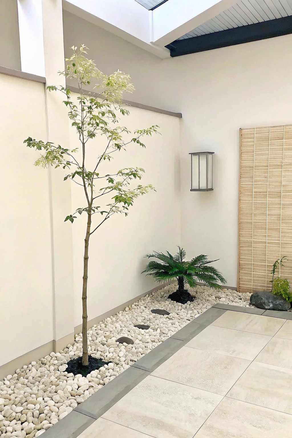 A narrow bed of white pebbles runs along a light wall next to a tiled patio, with a tall tree and small palm planted in the gravel.