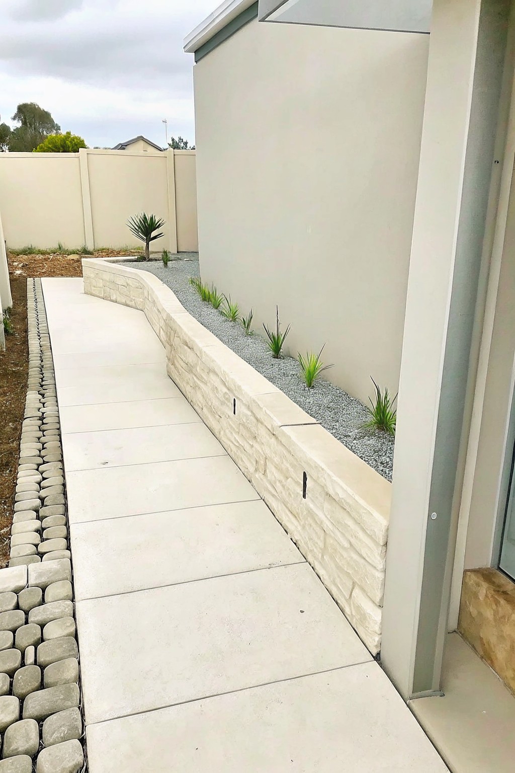 A concrete path runs beside a curved light stone retaining wall that borders a narrow gravel planting bed with small plants against a beige building wall.