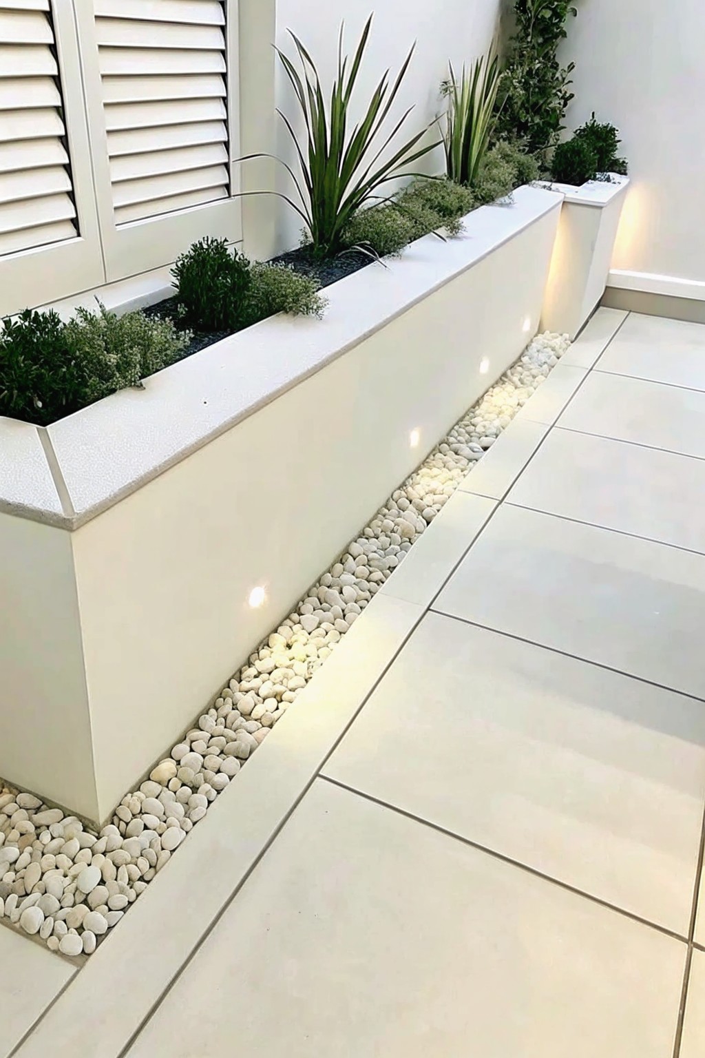 A long white raised planter filled with green plants runs along a tiled patio, separated by a strip of white pebbles with small embedded lights.