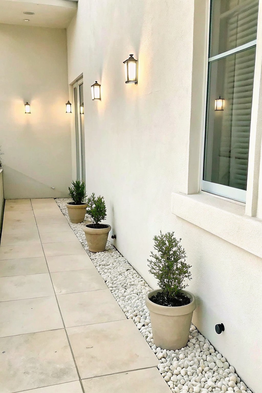 Lit lanterns illuminate potted shrubs on white pebbles along a tiled walkway.