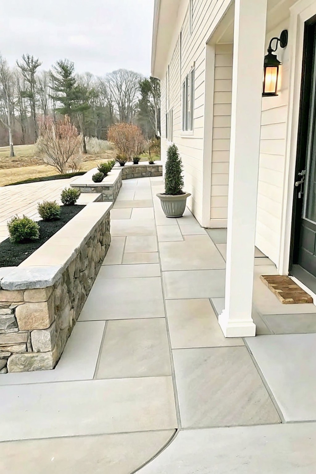 A wide stone pathway runs beside raised garden beds edged with stacked stone walls, planted with small shrubs in dark mulch, next to a house with light siding.