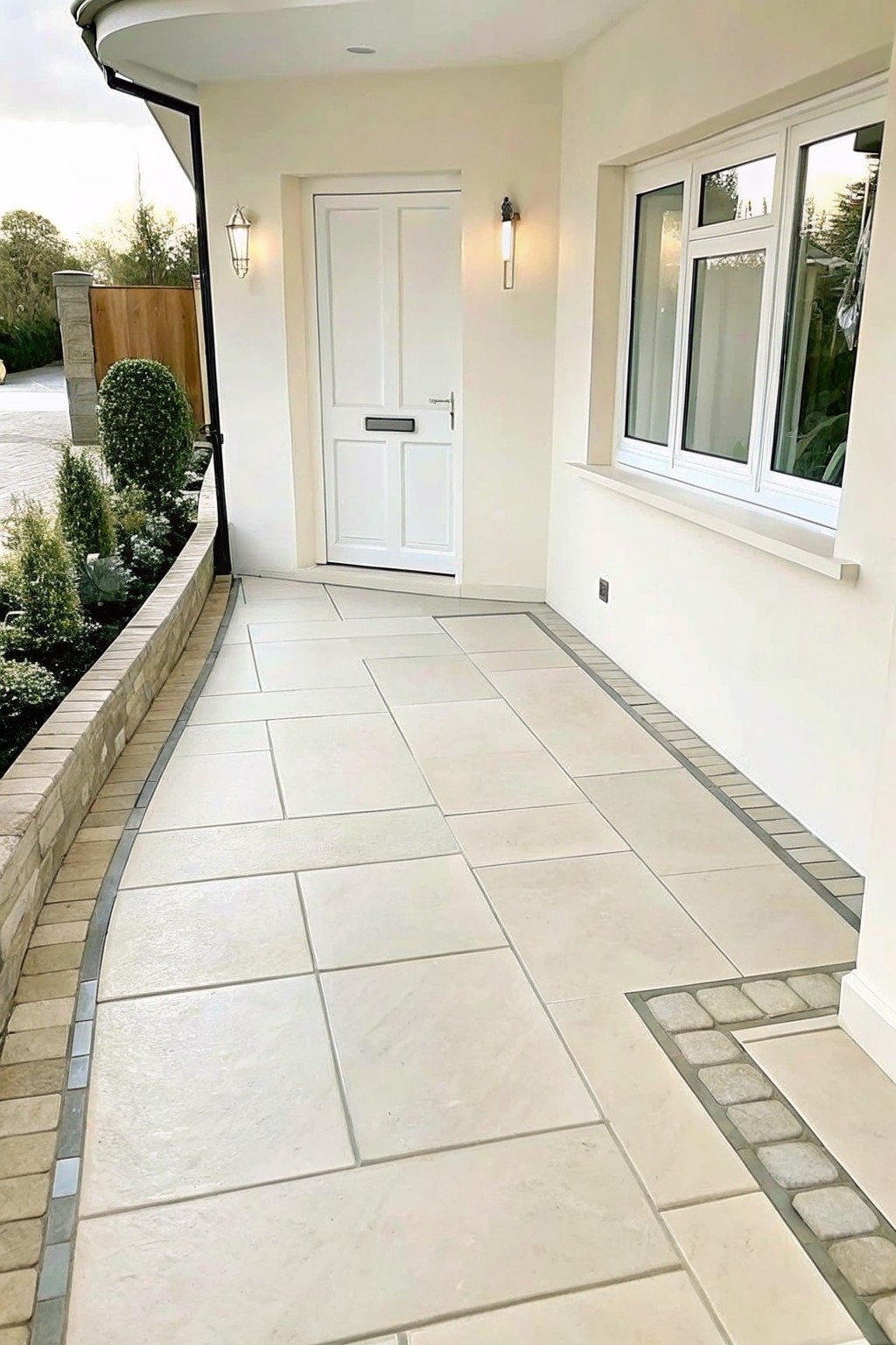 A light-colored paved walkway curves past a low stone border lined with shrubs and plants, leading toward a white front door.