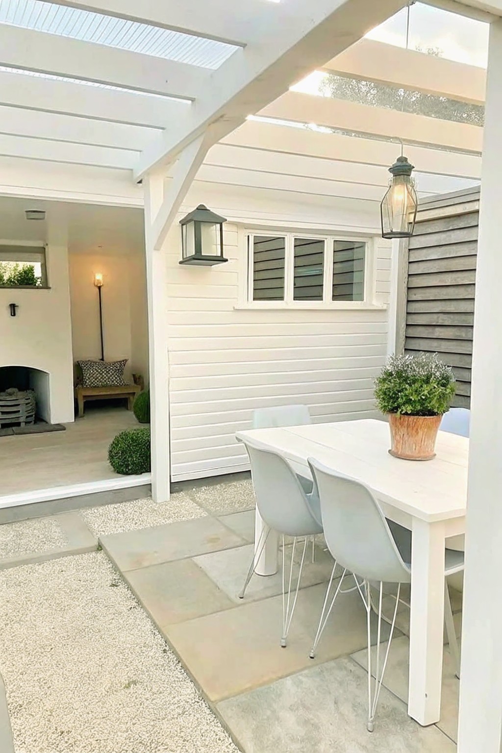 White patio table with gray chairs under pergola, lanterns, and potted plant on stone paving
