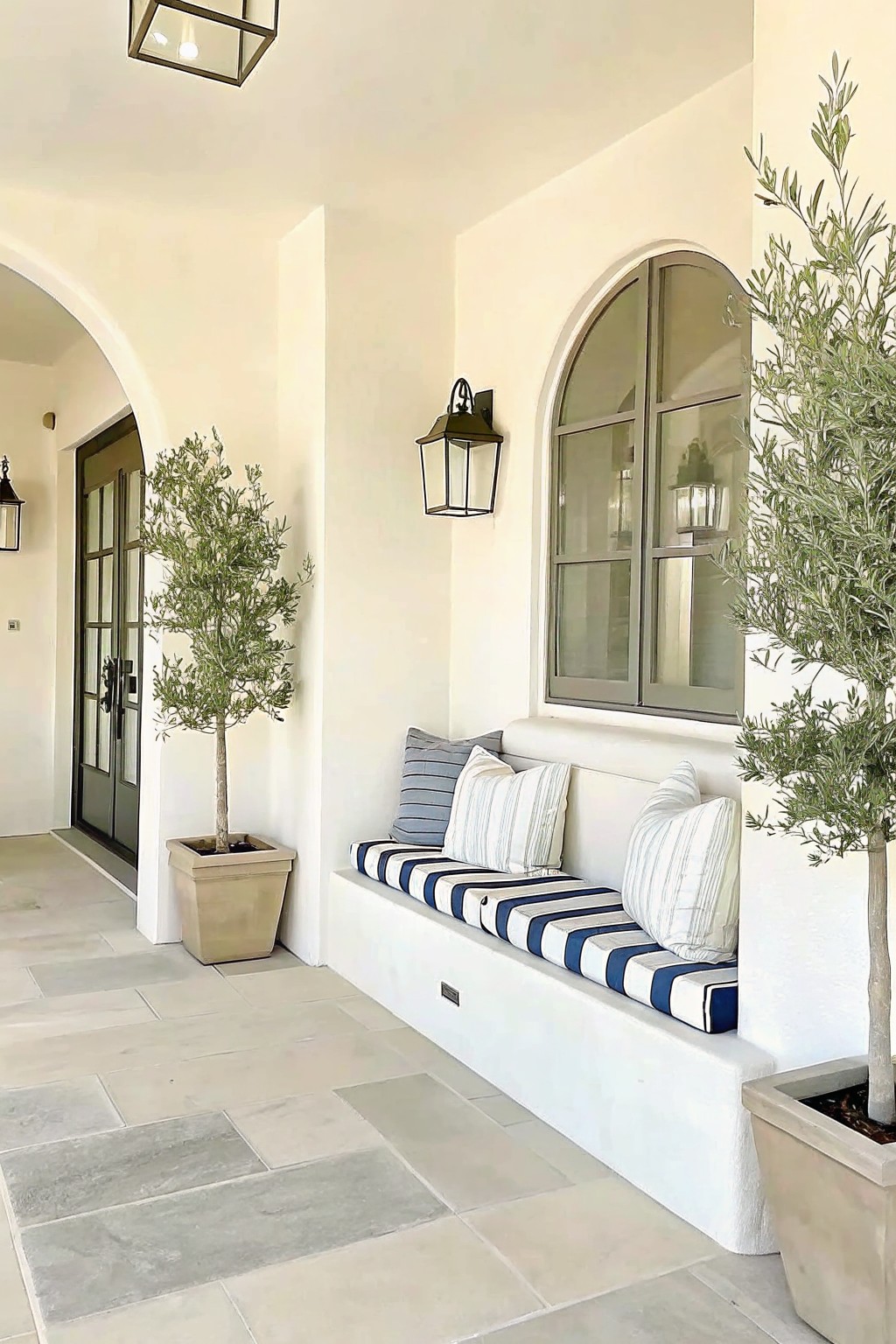 A covered patio with a white built-in bench topped with blue and white striped cushions, flanked by two large potted trees, next to an arched window and door.