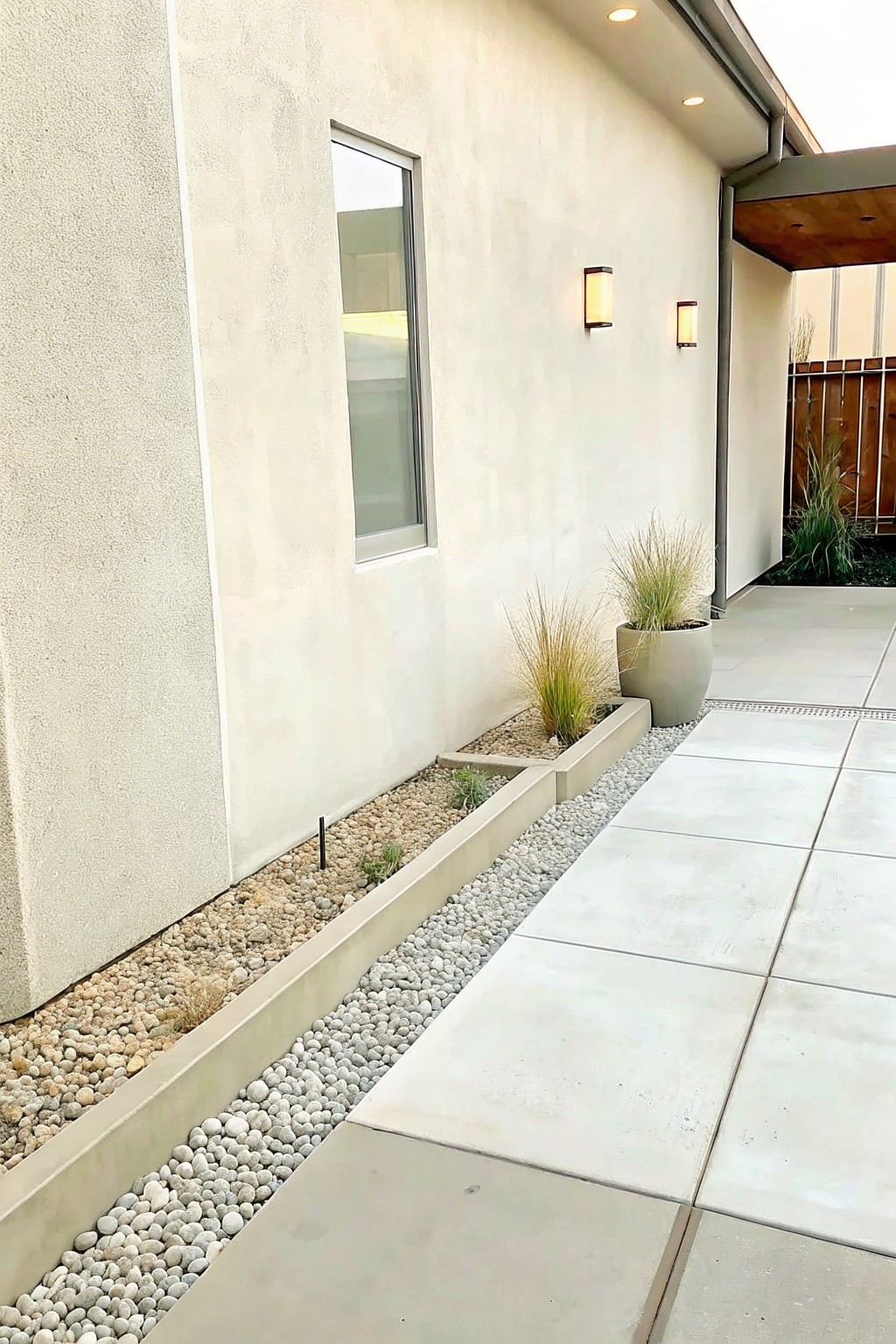 Modern stucco wall with narrow window, wall lights, pebble planters, and tiled patio.