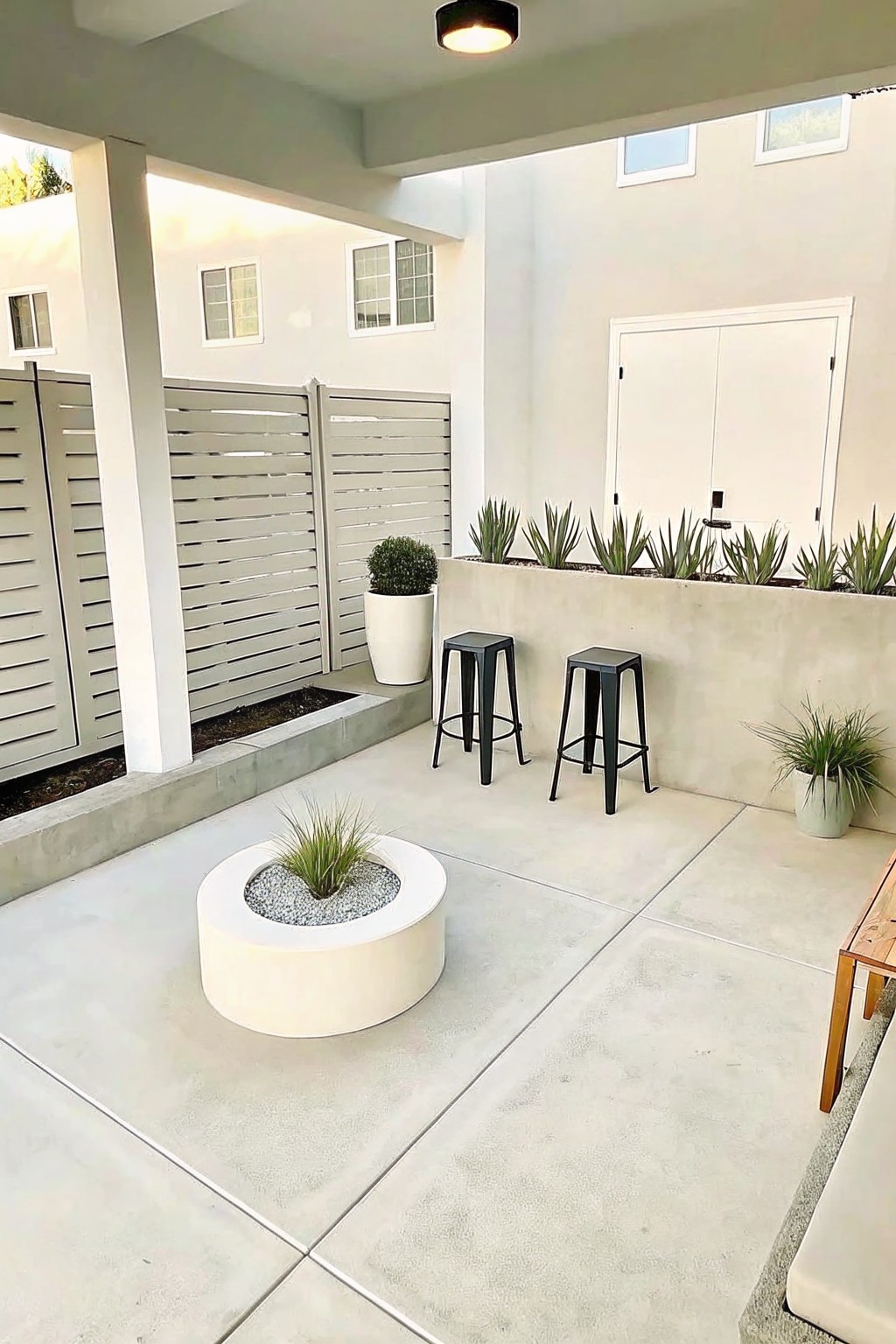 A concrete patio with a round central planter holding grass, two black stools, and a long raised bed planted with succulents along a wall.