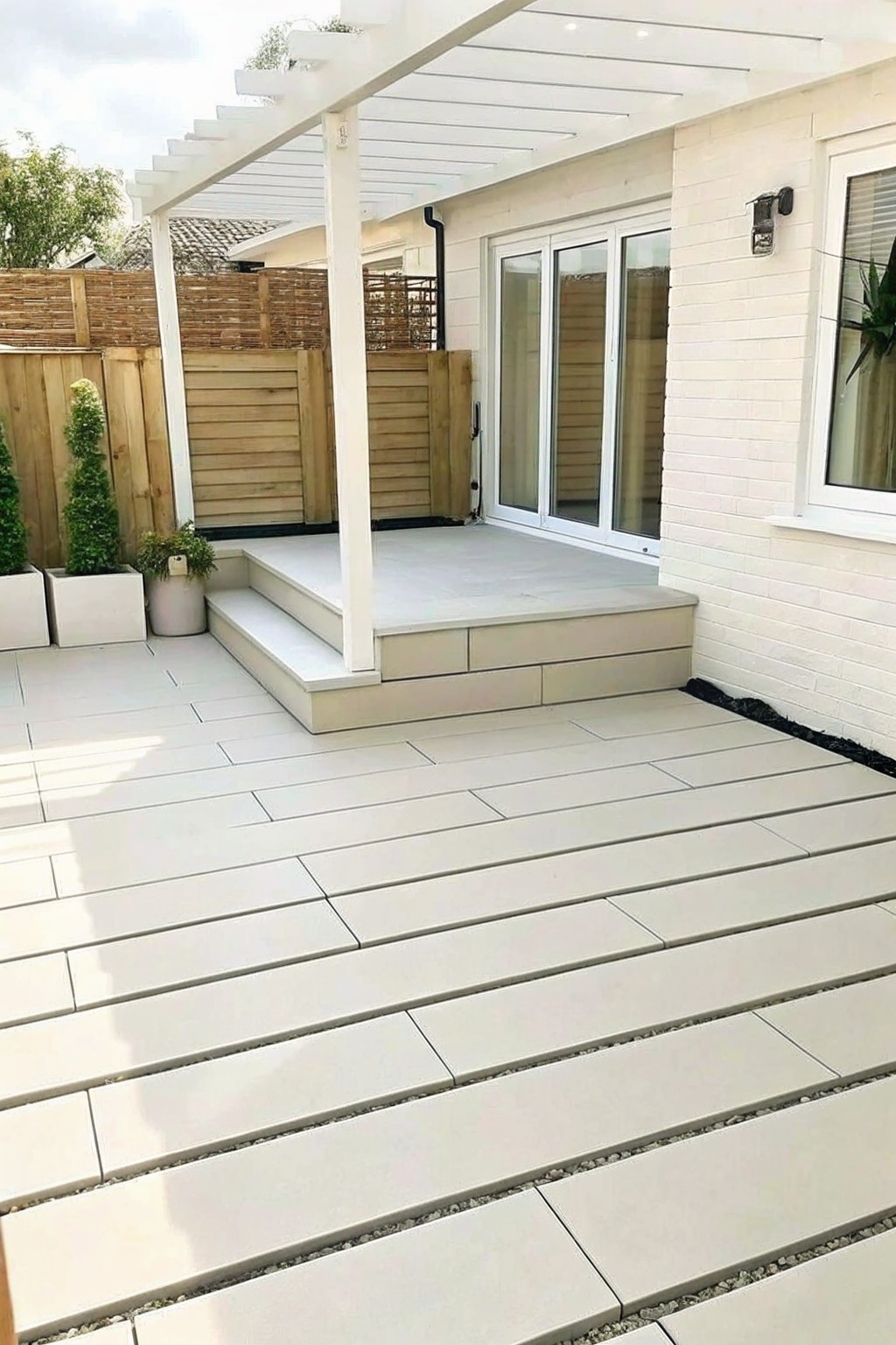 A backyard patio covered in large rectangular light gray pavers, with steps leading to a raised platform under a white pergola beside a white brick house.