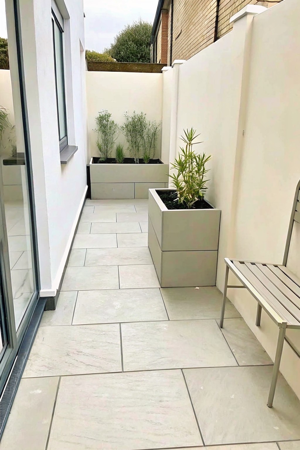 A narrow outdoor patio paved with light gray tiles features two large rectangular planters with green plants and a metal bench against the right wall.