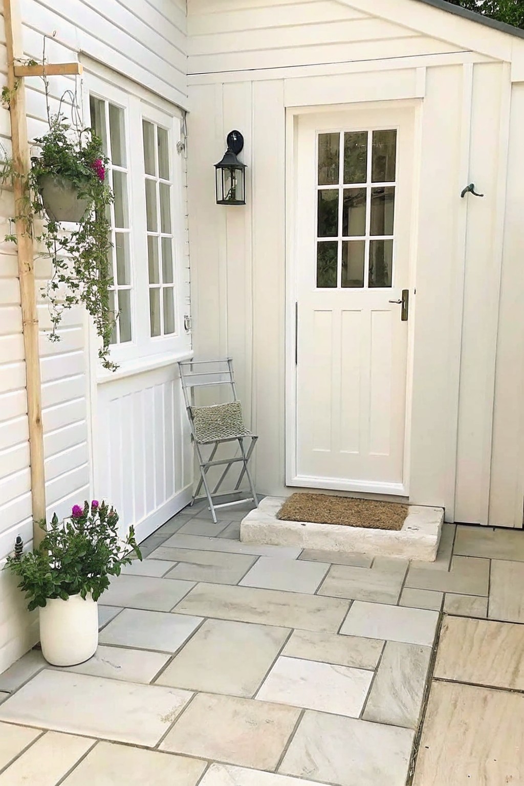 White house entry with paneled door, folding chair, lantern, and potted plants on patio.