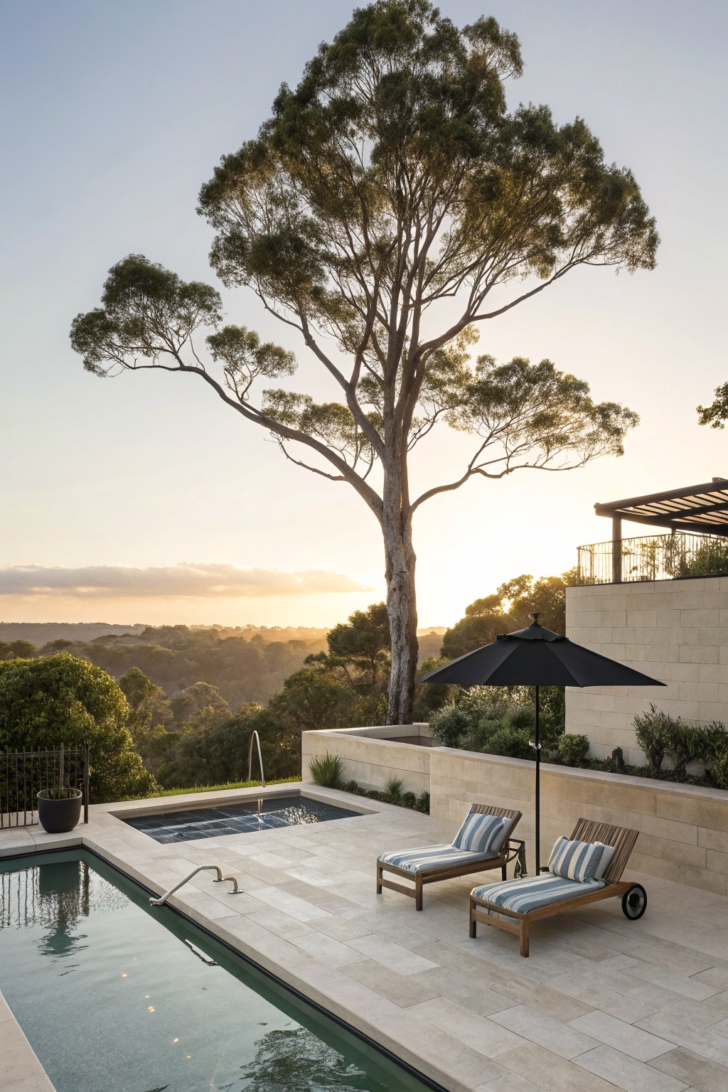 A pool deck with two lounge chairs under a black umbrella, a large tree behind the seating area, and a view of hills at sunset.