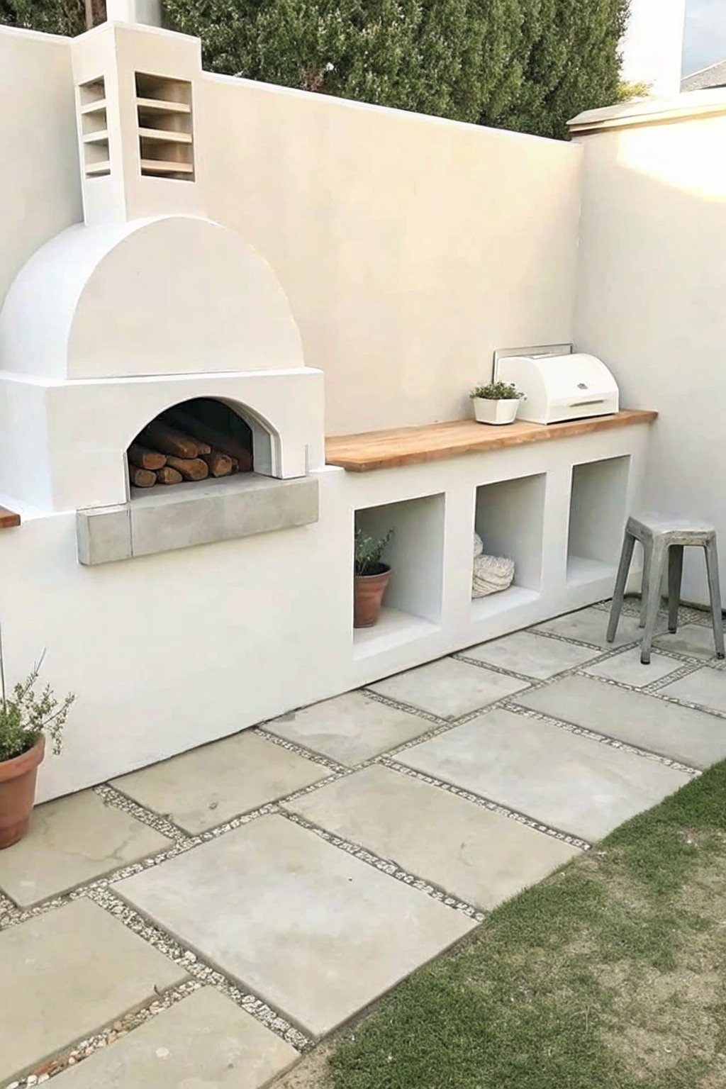 An outdoor pizza oven built into a white concrete structure with a wooden countertop and three open cubbies below, set on large stone pavers.