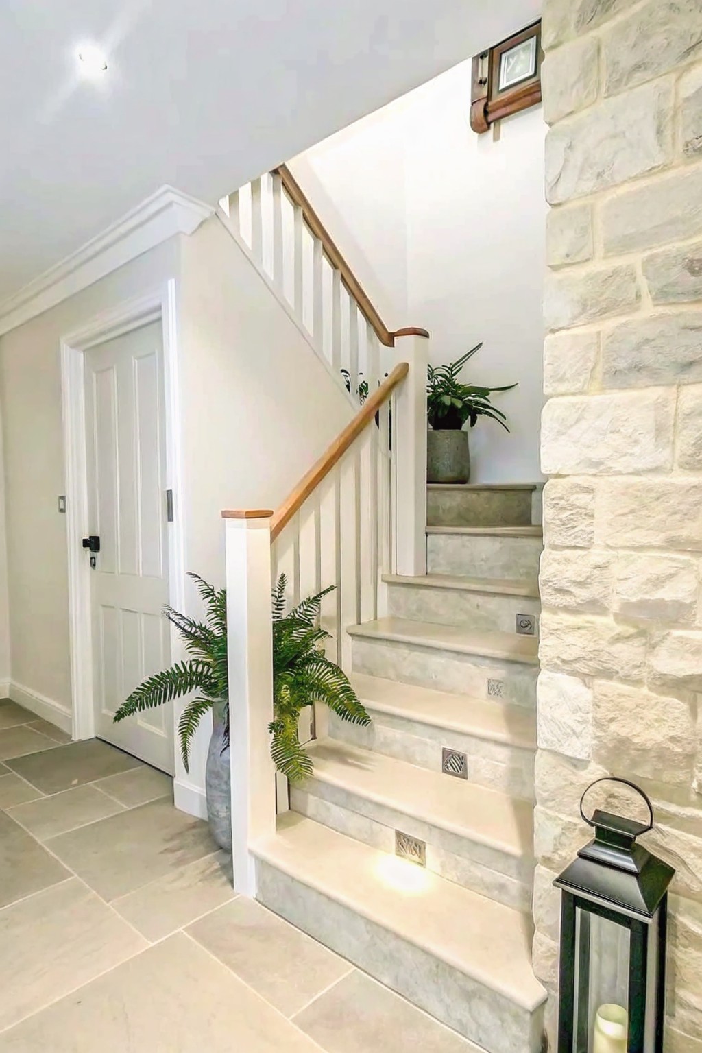 Interior staircase with a natural stone wall on the right, white balusters, wooden handrail, and potted plants at the base.
