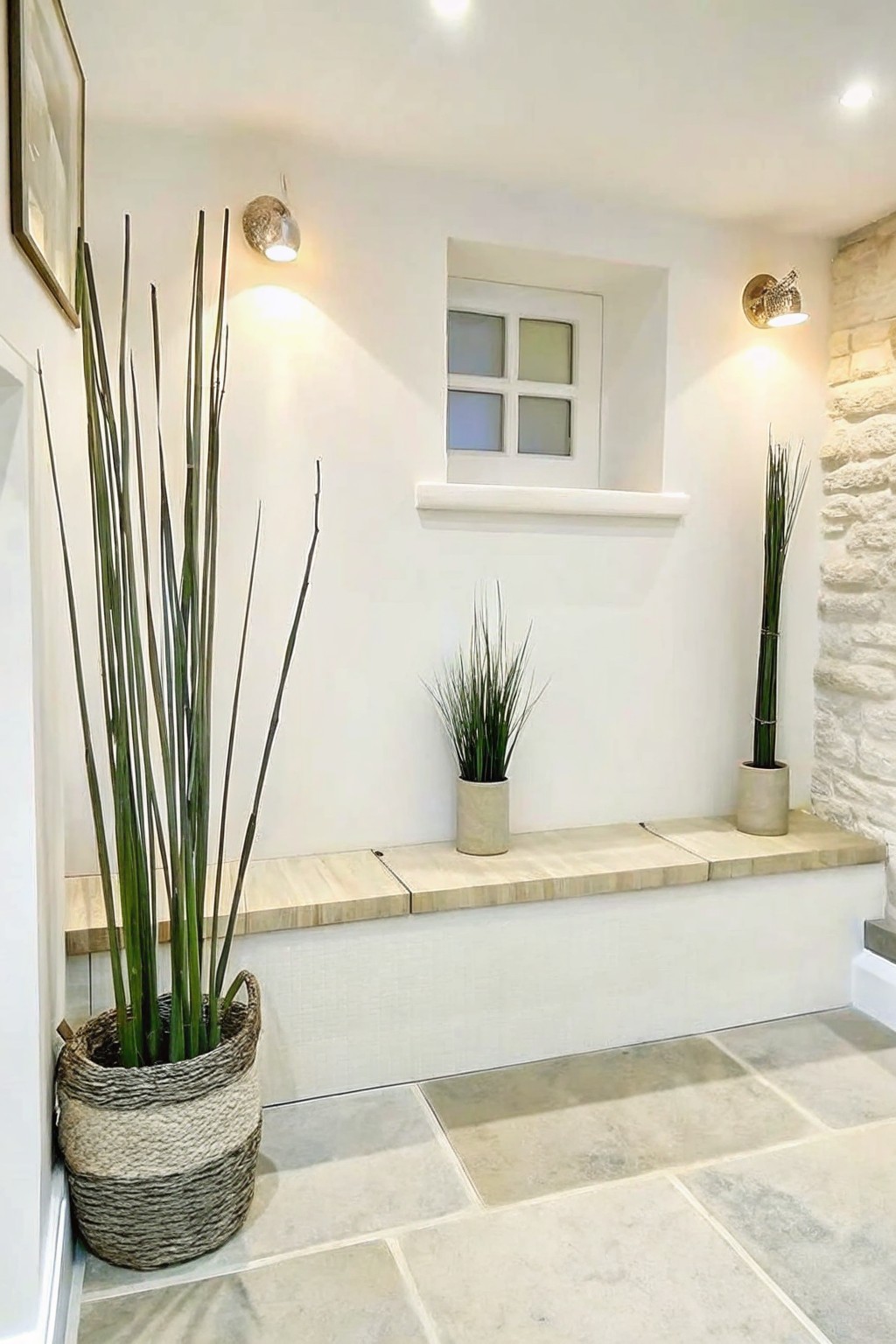 An indoor space with a long built-in bench of light stone slabs holding several potted plants, white walls, wall sconces, and a small window above the bench.