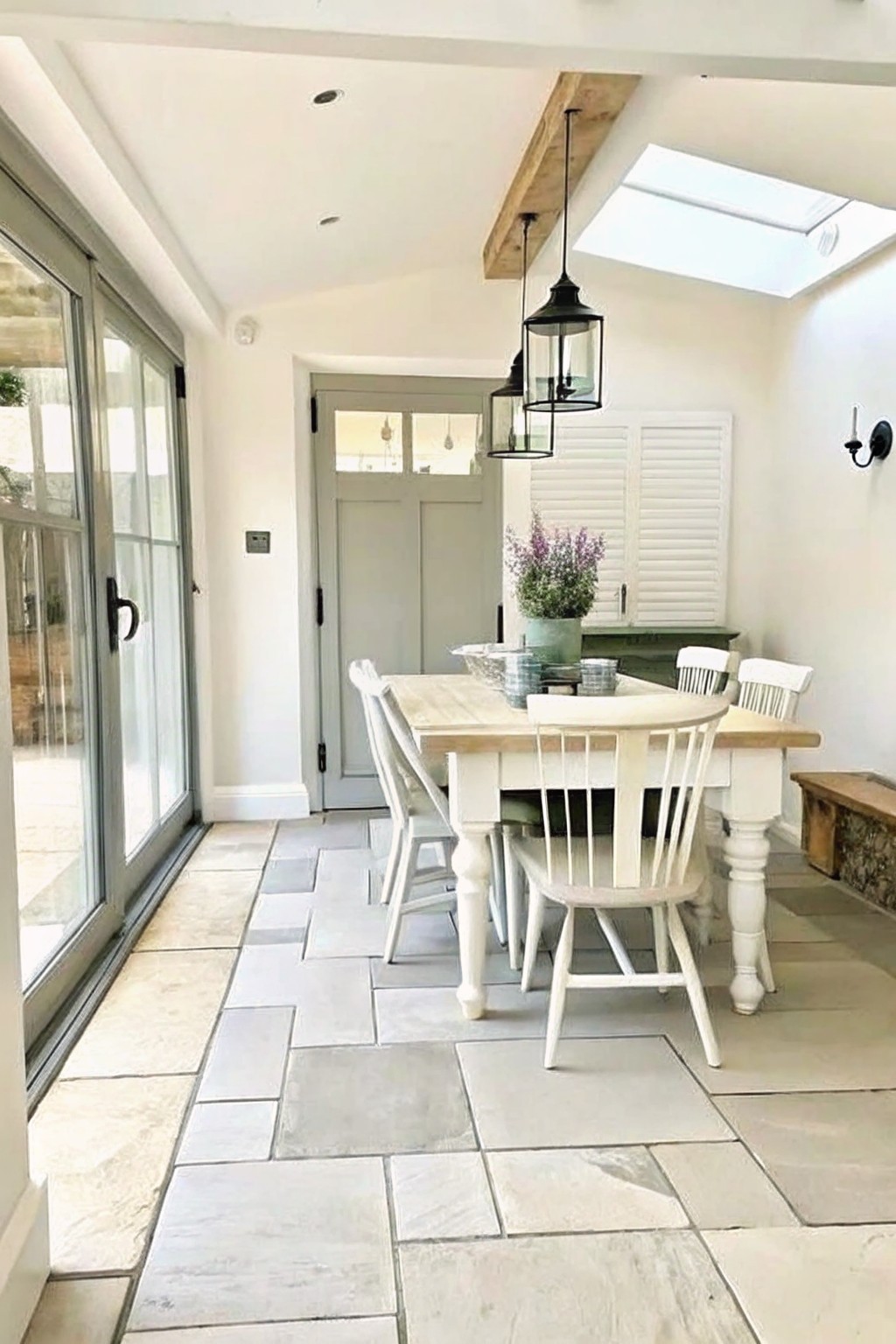 A bright dining room with a white wooden table and chairs set on large irregular gray stone tiles, a wooden ceiling beam overhead, and glass doors along one wall.