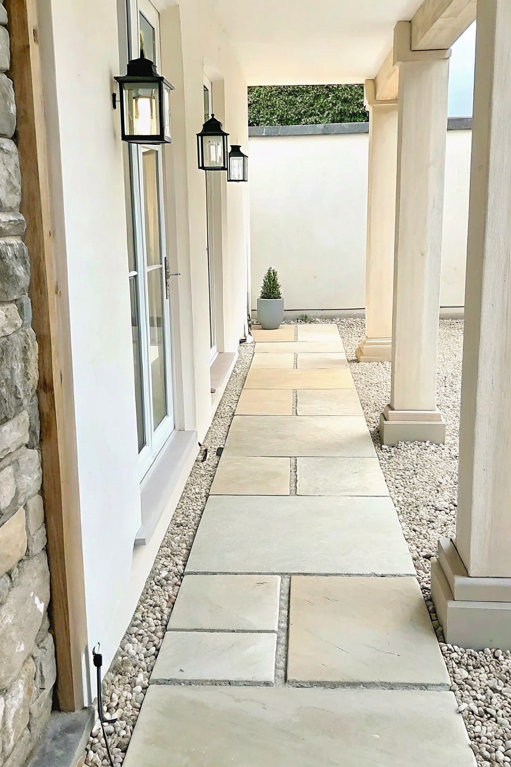 A straight garden path of large rectangular stone pavers bordered by light gravel, running beside a white wall with black lanterns and a small potted shrub in the distance.