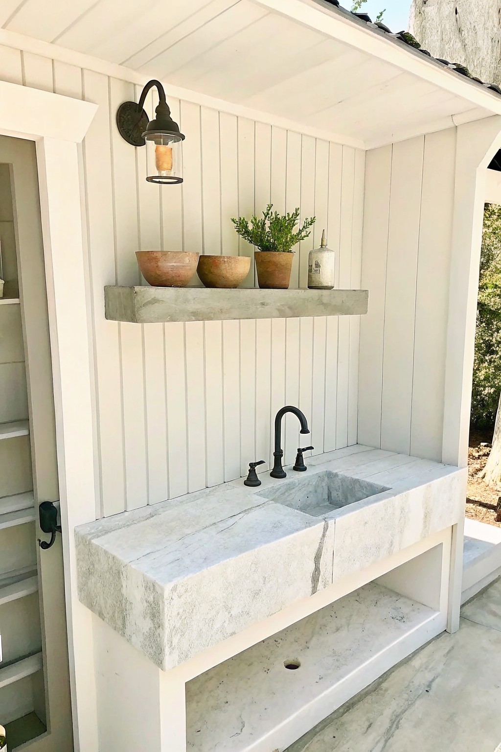 An outdoor corner with white wood walls holds a large natural stone sink with black faucet and a floating concrete shelf above it.