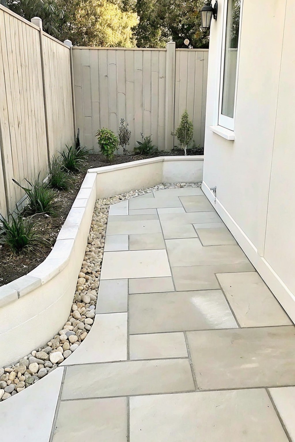 A garden path of large light-colored stone pavers bordered by gravel and low plants along a wooden fence.
