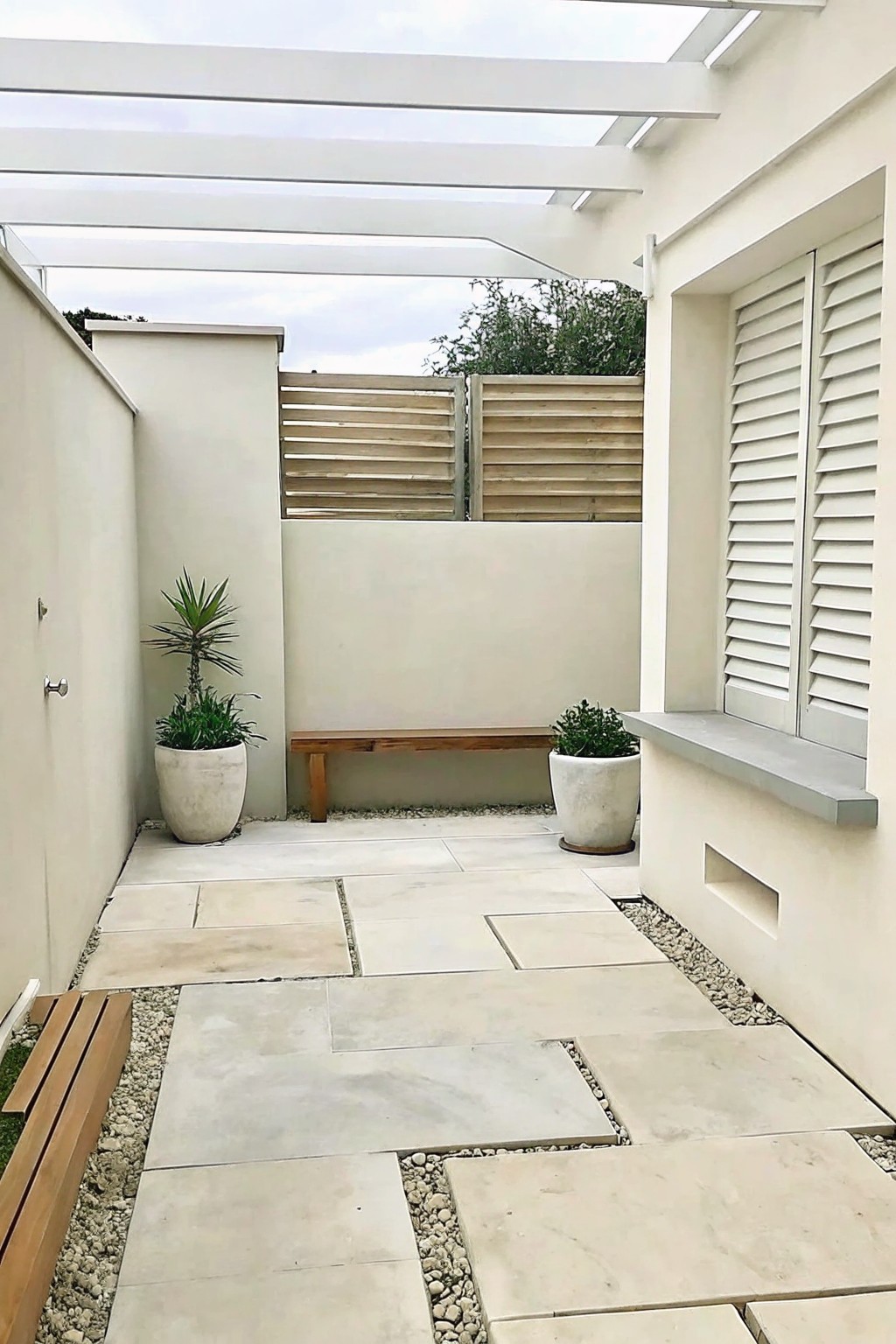 A small courtyard patio paved with large light-colored stone slabs set in gravel, featuring two wooden benches, potted plants, white walls with shutters, and a white overhead pergola.