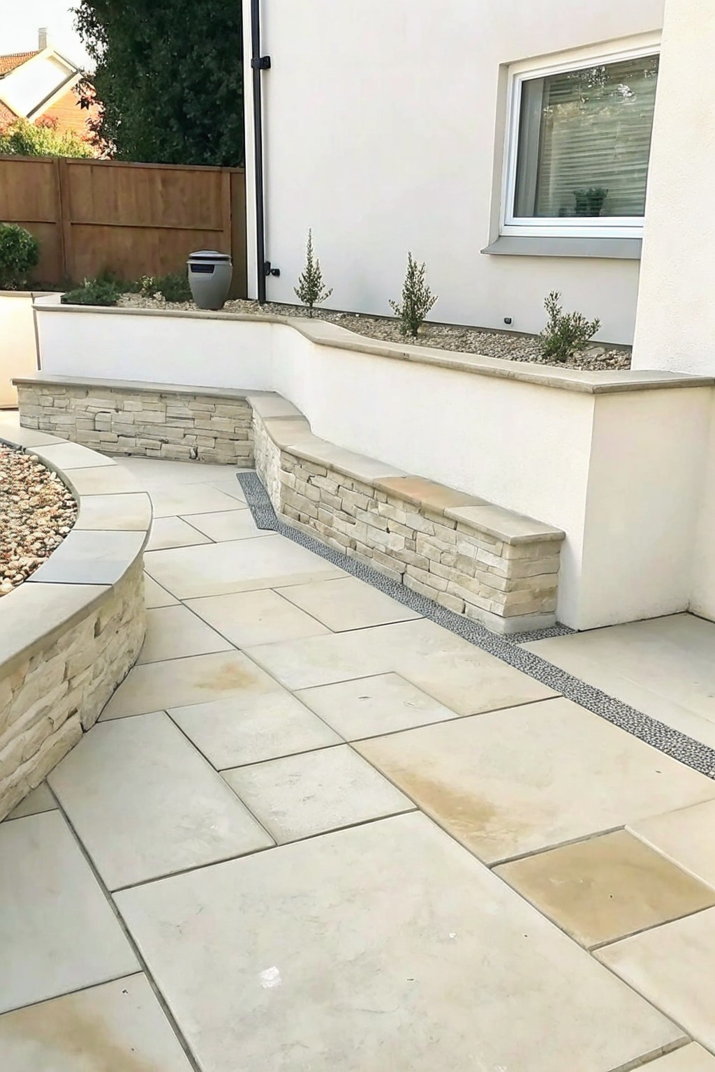 A patio paved with large light stone tiles features curved low walls of stacked natural stone with flat caps used as seating, alongside raised beds filled with gravel and small shrubs next to a white house.