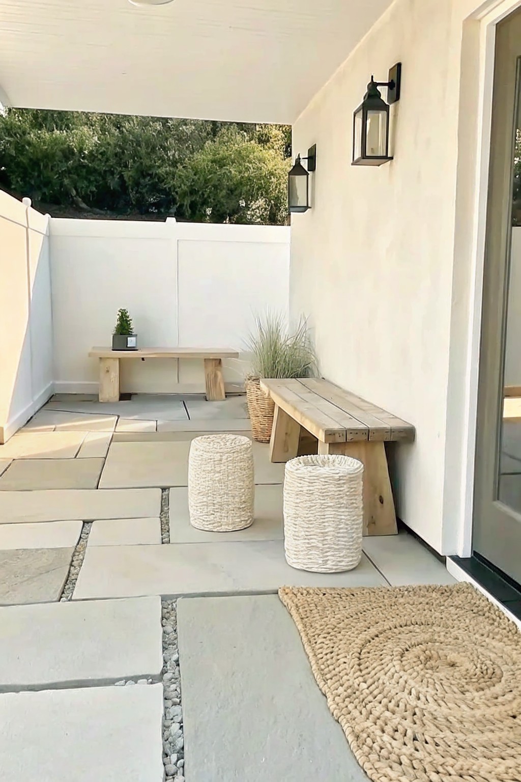Outdoor patio with wooden benches, woven stools, lanterns on white wall, jute rug