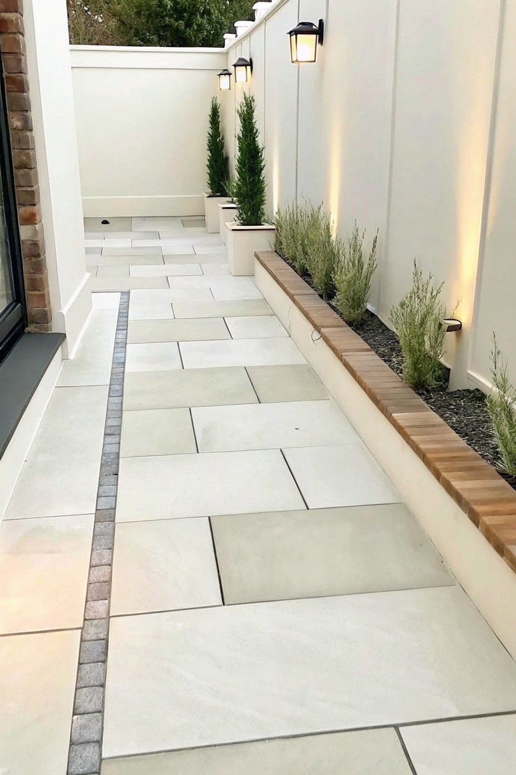 A narrow paved walkway runs beside a white wall with a long raised garden bed edged in brick and planted with low shrubs and taller evergreens.