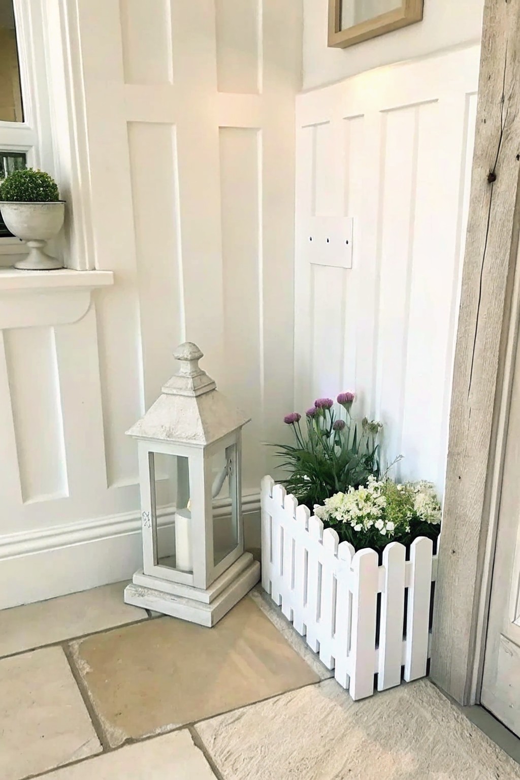 White lantern beside picket fence planter with purple and white flowers on stone floor.