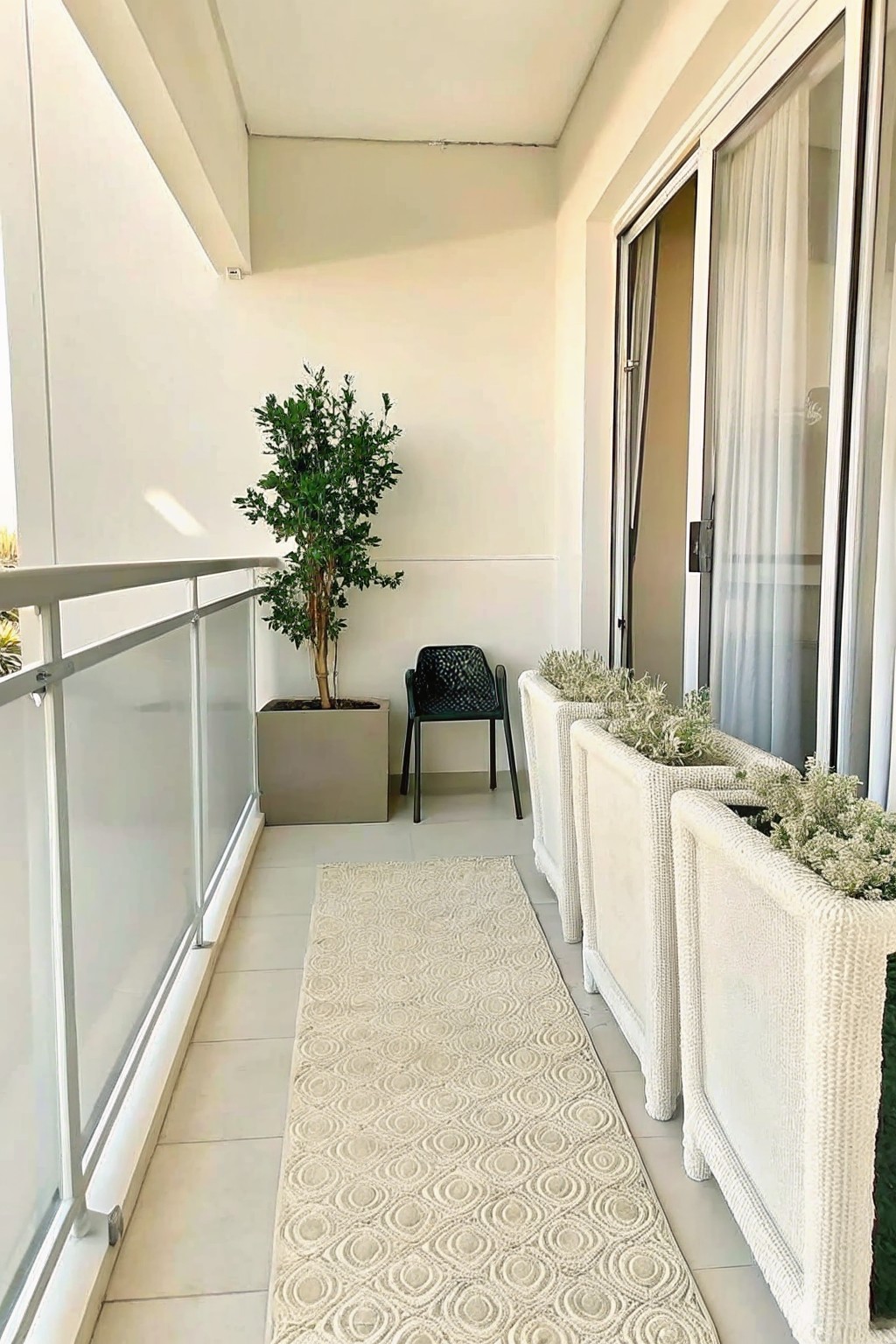 A narrow balcony features a row of white rectangular planters with greenery along the right side, a large potted tree in a gray container, a black chair, and a patterned runner on the floor.