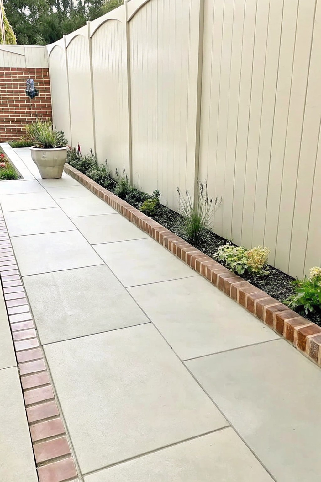 A long concrete path edged with a single row of red bricks bordering a mulched flower bed with low plants against a tall beige fence.