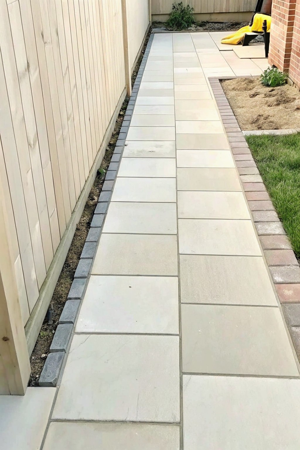 A straight paved garden path with light-colored stones bordered by dark gray blocks on the left and multicolored bricks on the right next to grass and a planting bed.