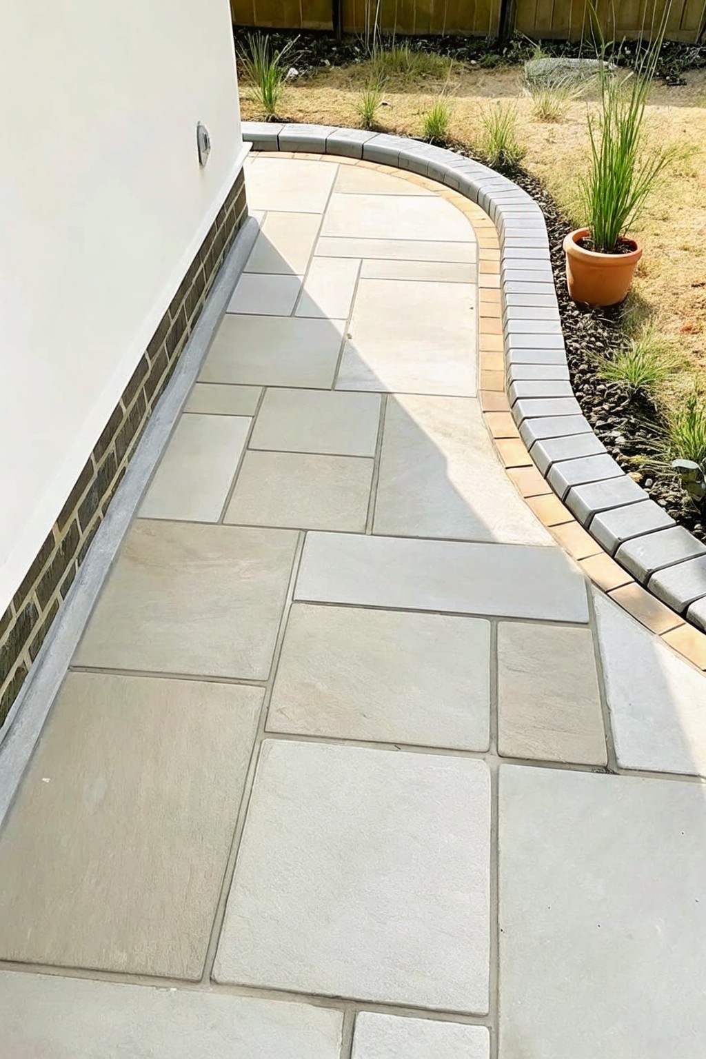 A curved brick border runs alongside a stone paved walkway next to a garden bed with mulch and plants.