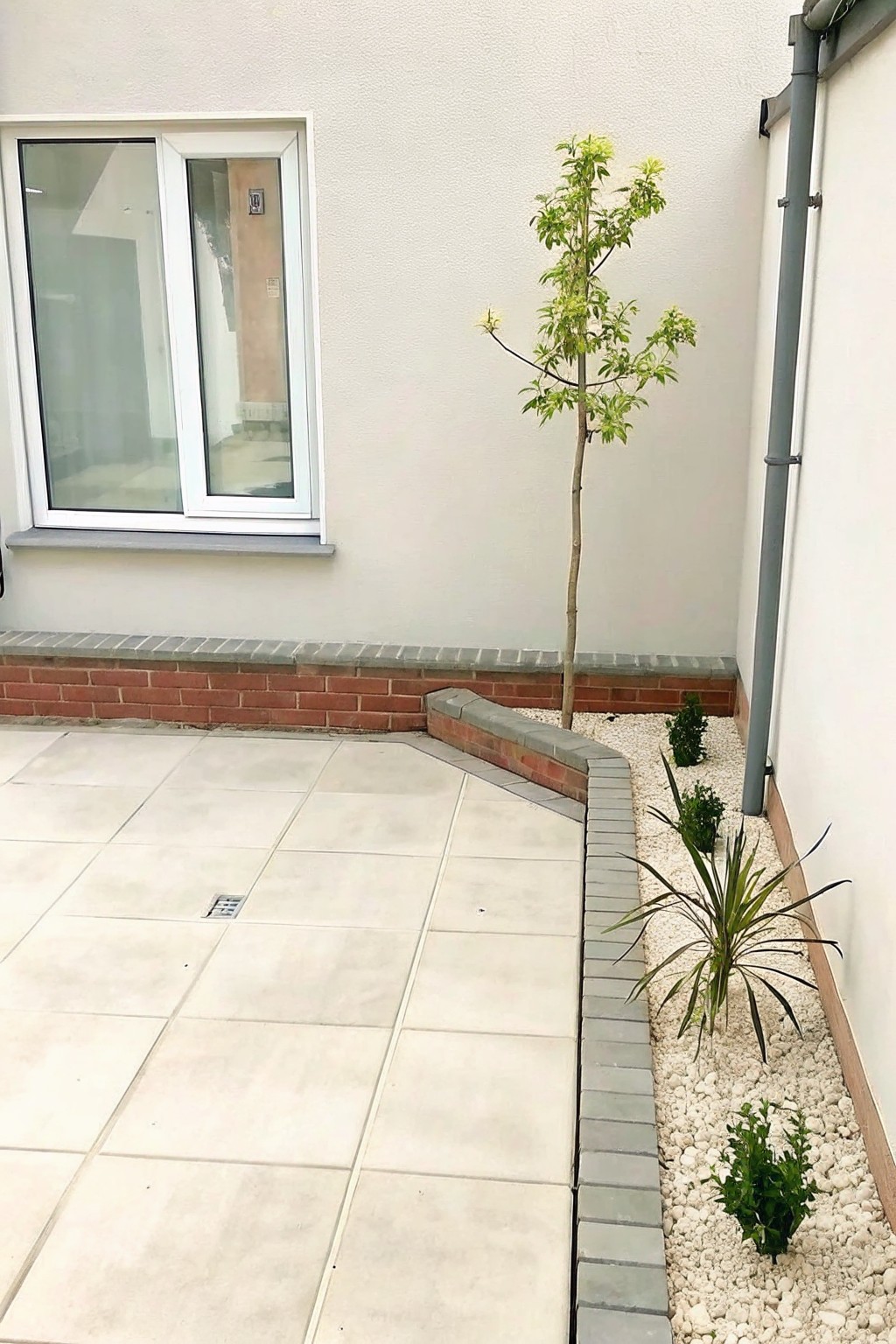 Patio with beige tiles, young tree in pebble bed by white wall with window