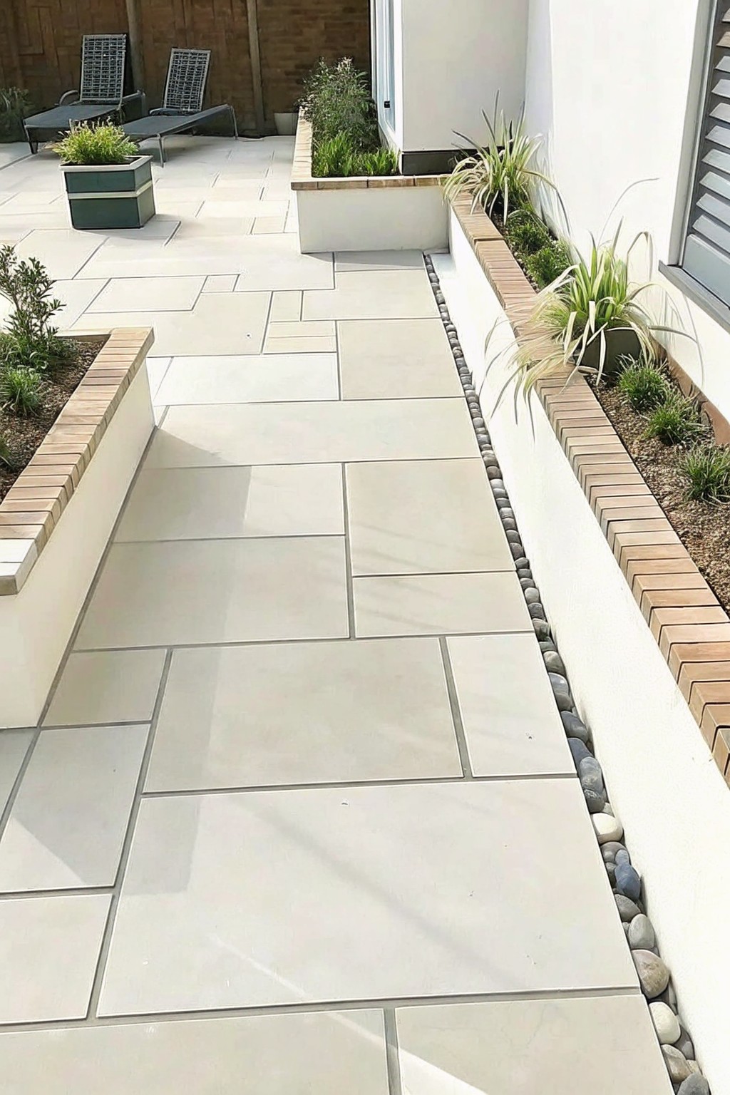 A light-colored paved walkway lined with low brick-edged garden beds containing plants and pebbles.