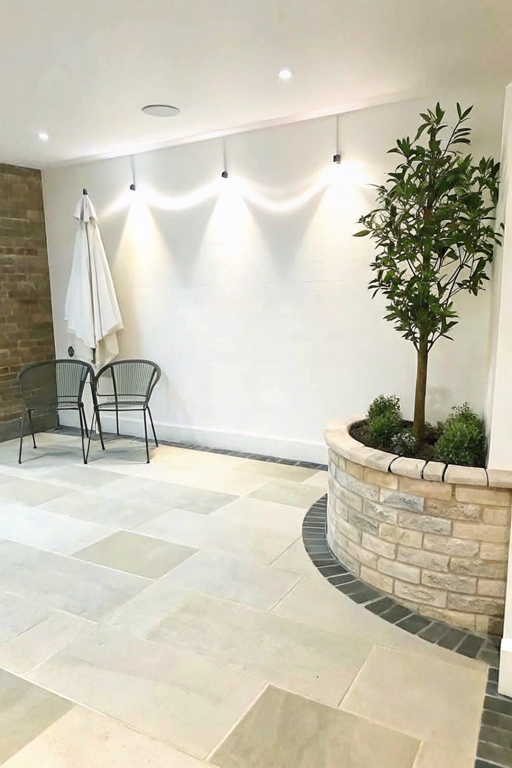 An outdoor patio with a curved brick planter holding a tall tree and smaller shrubs beside a white wall and two metal chairs.