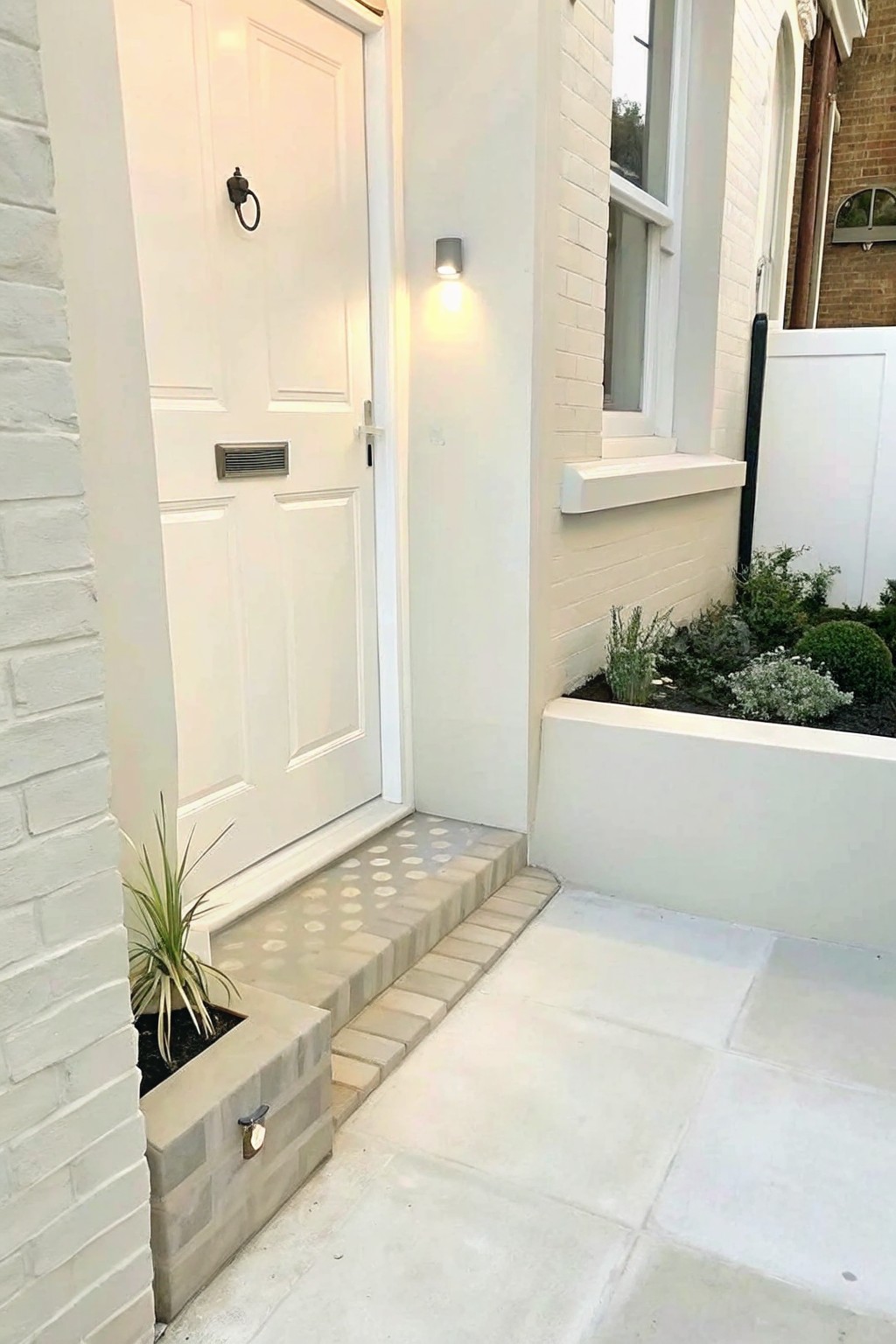 A white front door with steps leads to a small raised concrete block planter holding a single plant, next to a larger garden bed along a white brick wall.