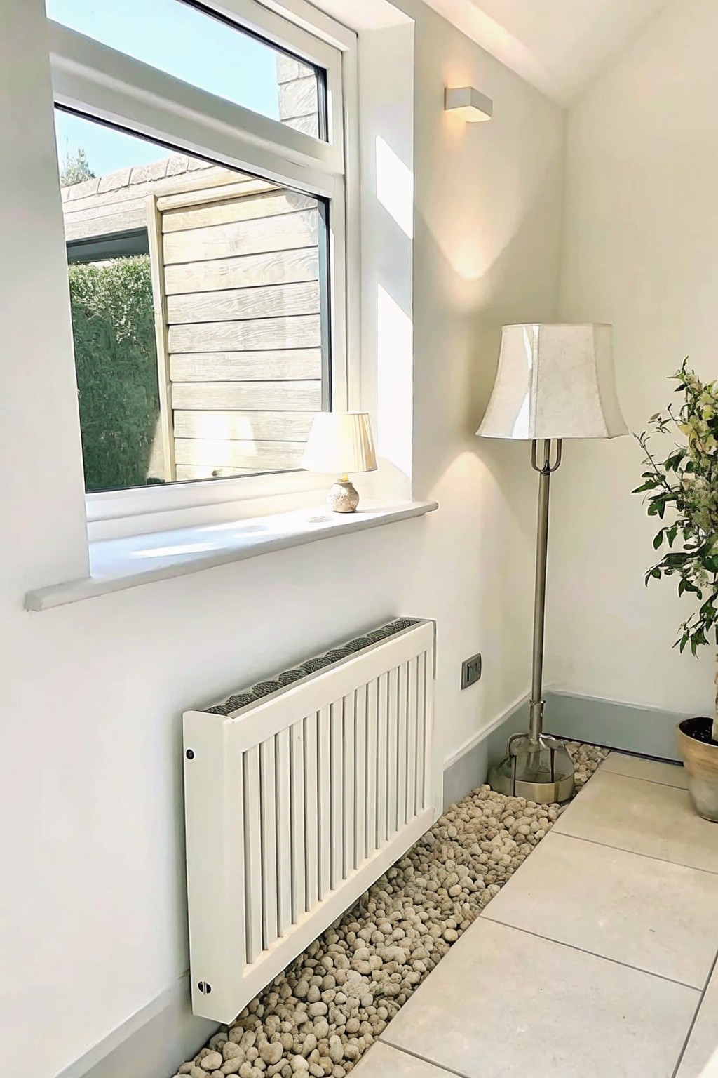 A white radiator sits above a bed of light-colored pebbles on a tiled floor next to a window with a floor lamp and potted plant nearby.