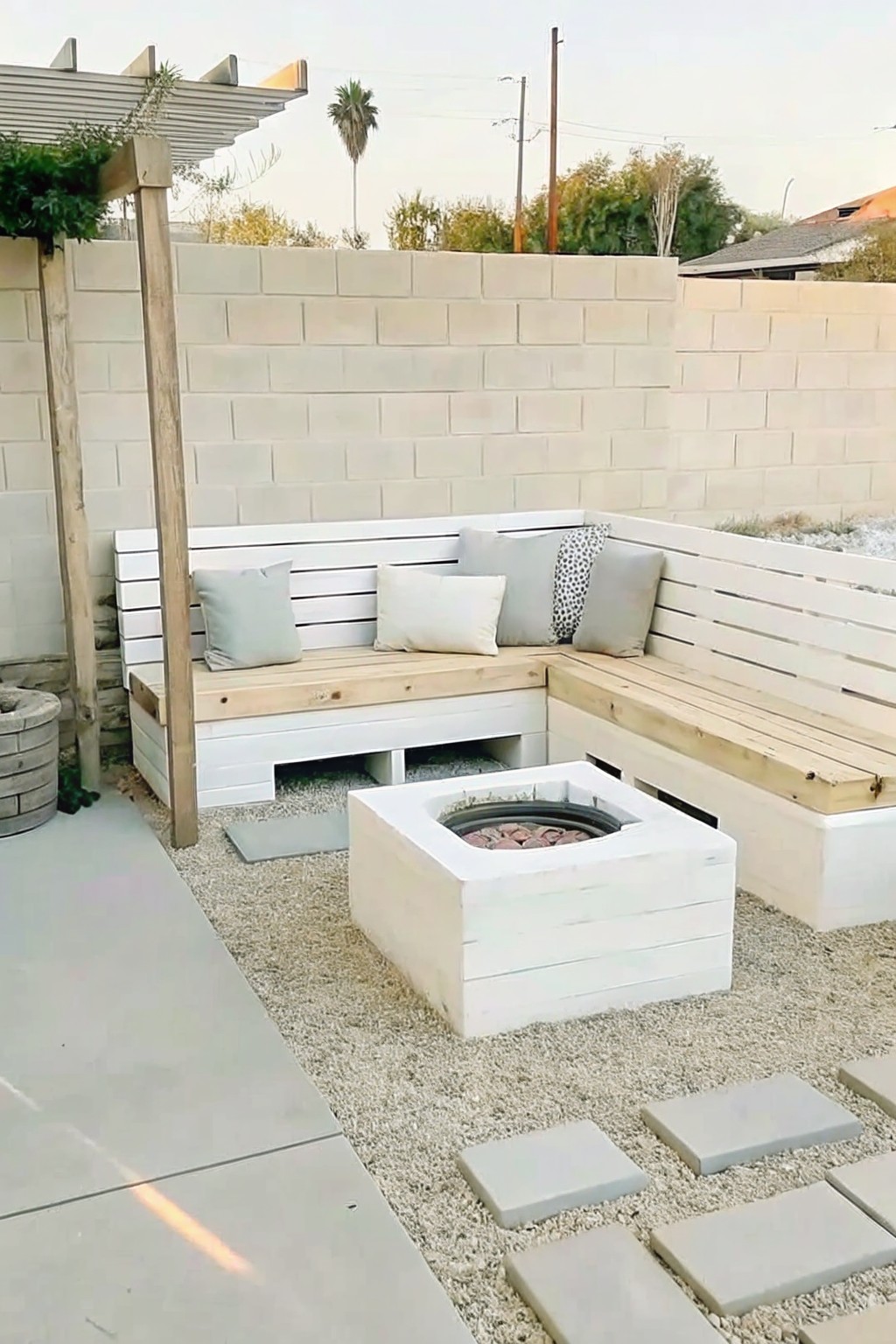 An L-shaped white wooden bench setup surrounds a square fire pit on gravel, backed by a cinder block wall and a wooden pergola.
