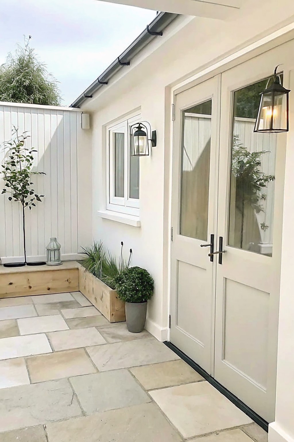 White double doors open onto a stone-paved patio lined with long wooden planter boxes holding shrubs and grasses, with wall lanterns beside the doors.