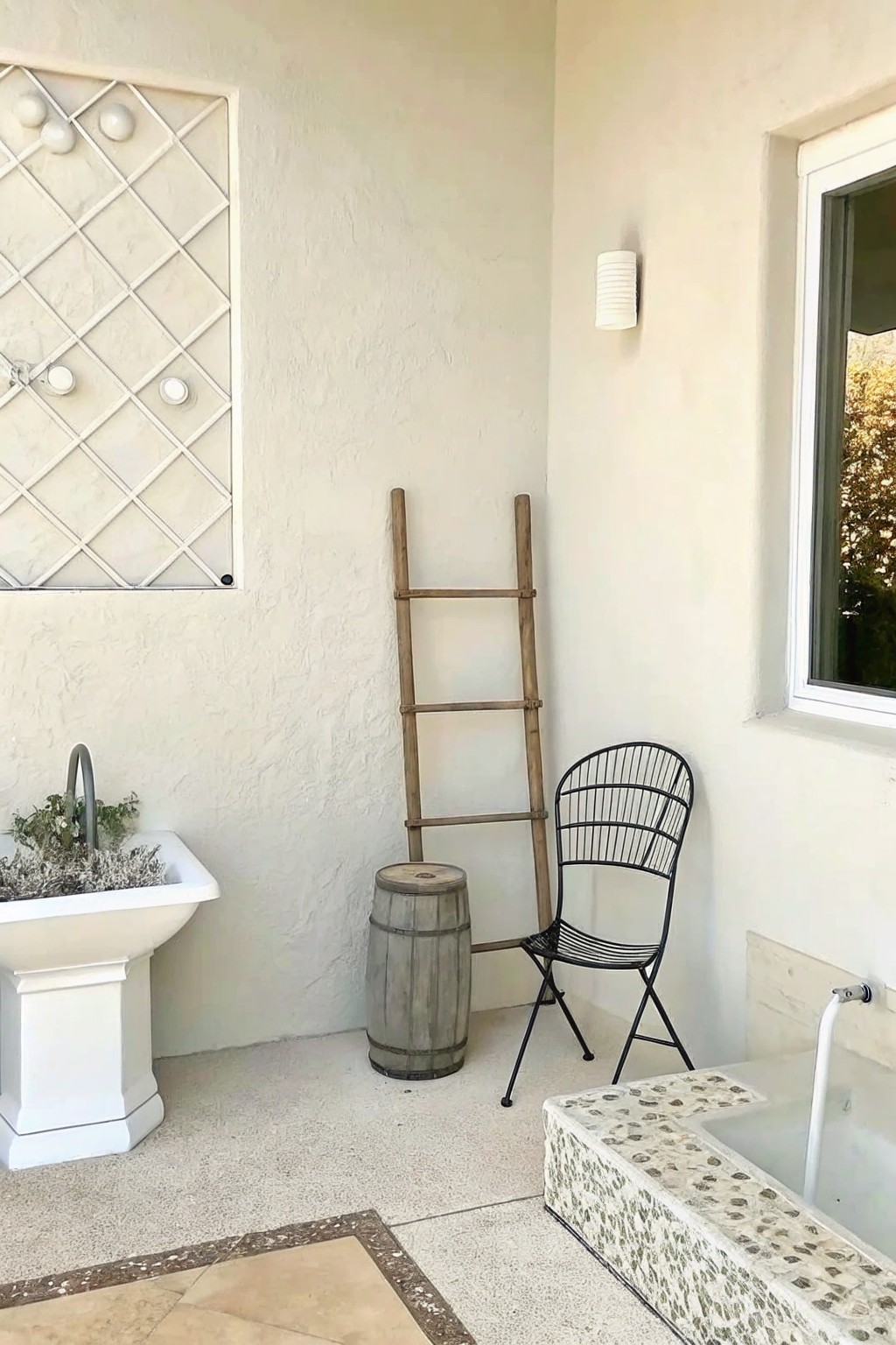A weathered wooden ladder leans against a stucco wall in an outdoor patio area beside a wooden barrel and black metal chair.