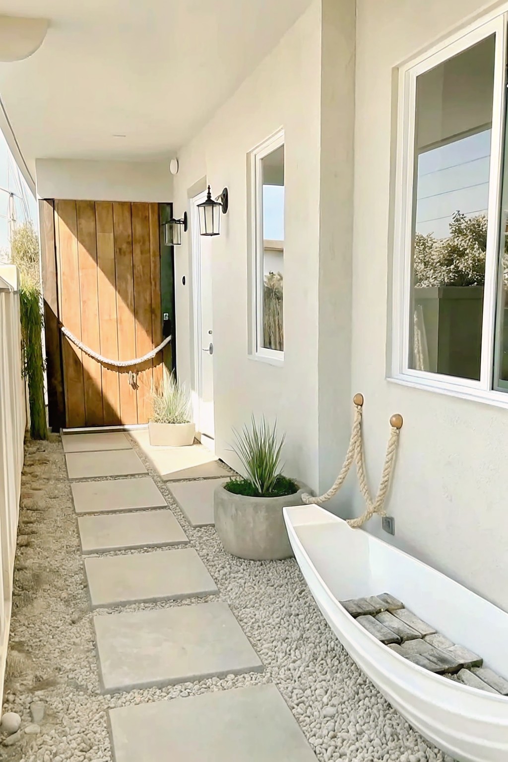 A narrow side yard path made of square pavers on gravel leads past potted plants to a wooden door, with a white boat repurposed as a planter resting against the house wall.