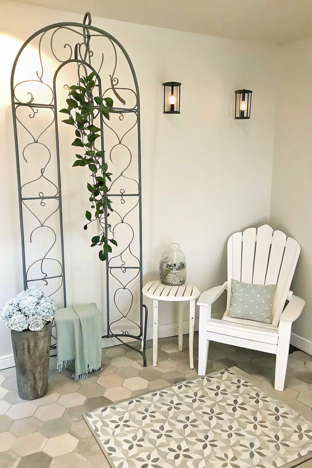 An indoor corner featuring a tall metal trellis with a climbing plant, a white Adirondack chair, a small side table, wall lanterns, and a vase of flowers on a hexagonal tiled floor.